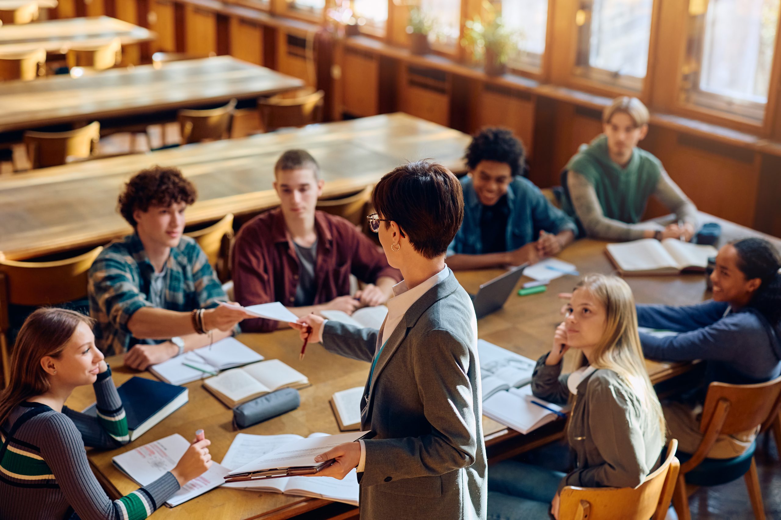 Female professor giving exam papers to group of high school students in the classroom.